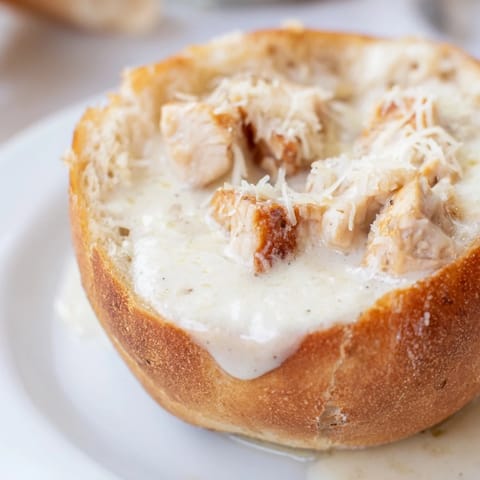 Four serving-sized crusty bread bowls, each filled with a rich and luscious chicken Alfredo pasta dish, garnished with fresh parsley.